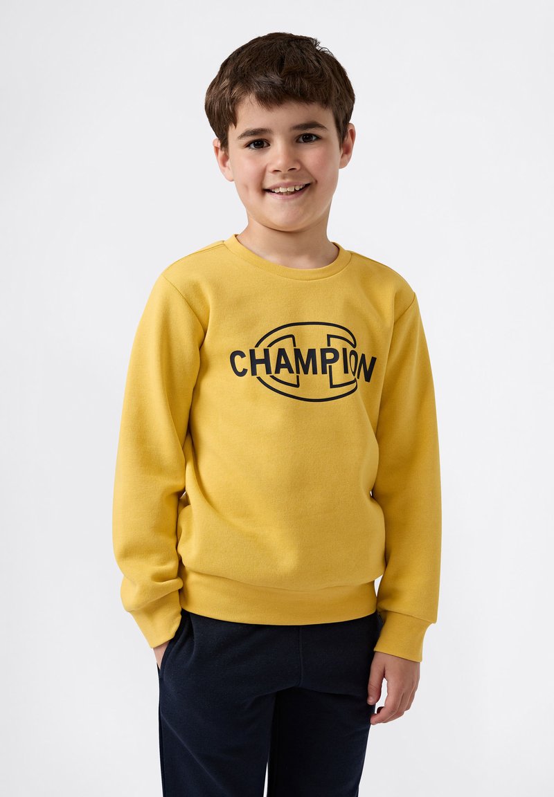 Geel sweatshirt gemaakt van zachte stof met een ronde hals, voorzien van een zwart "CHAMPION" logo op de borst. Draagt met donkere broeken.