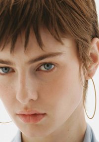 Pendientes de aro dorados, de tamaño mediano, con un acabado suave. Cabello corto y texturizado de color marrón y piel clara con un maquillaje mínimo. Enfoque en las características faciales.