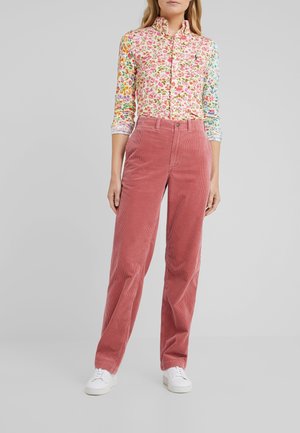 Woman wearing a floral long-sleeve button-up shirt, high-waisted dusty pink corduroy pants, and white sneakers, standing against a gray background.