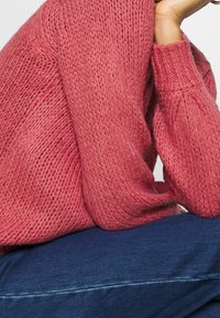 Silný pletený svetr v jemné rů�žové barvě. Má volný střih, žebrované manžety a texturovaný povrch. Kombinován s tmavě modrým denimem.