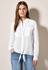 Camicia bianca con bottoni realizzata in tessuto leggero, con due tasche sul petto, maniche lunghe e orlo annodato. Indossata con jeans blu.