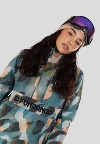 Mladá žena s dlouhými tmavými vlasy má na sobě lyžařskou bundu s multicolor maskáčovým vzorem, růžovou čepici a fialové lyžařské brýle na hlavě.