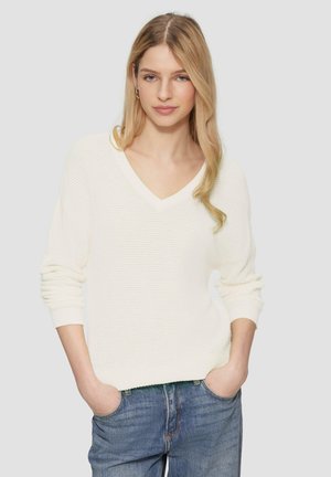 Weißer Strickpullover mit V-Ausschnitt und langen Ärmeln, aus strukturiertem Material mit gerippten Bündchen, getragen über blauen Jeans.