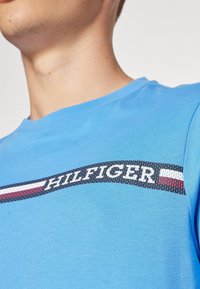 Ljust blå bomullströja med en grafisk band som har "HILFIGER"-text och röda och vita ränder, rund hals och korta ärmar.