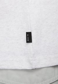 Vit, texturerad t-shirt i tyg med en synlig svart etikett som läser "MAX 90" nära fållen, lager på ett grått tyg. Enkel design.