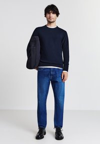 Sweatshirt marine à col rond, col intérieur gris clair, jeans bleus et chaussures noires ; modèle tenant un grand sac fourre-tout noir.