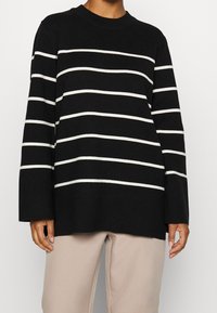 Maglione a righe nere con righe orizzontali bianche, scollatura rotonda e maniche lunghe, abbinato a pantaloni beige chiaro, texture morbida.