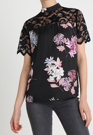 Schwarze Bluse mit floralen Mustern in Pink und Blau; Spitzenärmel und hoher Kragen, kombiniert glattes Gewebe mit strukturierten Akzenten.