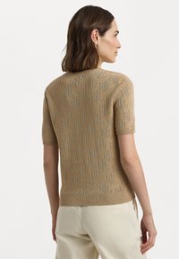Beige īsām piedurknēm adīta džempera ar teksturētu saistīšanu, kas iezīmē smalku visapkārt esošo logotipu rakstu. Ribota apkakle un apakšmalā noslēdz dizainu.