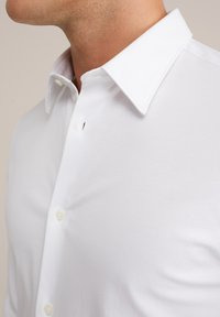 Man in een frisse witte blouse met een puntkraag, tegen een neutrale achtergrond, goed zichtbaar zijn hals en bovenborst.