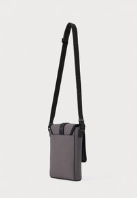 Compatta borsa a tracolla verticale grigia con tracolla nera regolabile e chiusura a patta su sfondo bianco.