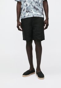 Calções pretos de linho com um corte descontraído, combinados com uma camisa de manga curta com estampa abstrata azul e cinza e espadrilhas pretas.