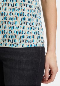 Bavlněný top s geometrickým modro-beige vzorem, s rounded lemováním a malou značkovou etiketou na bočním švu, spárovaný s tmavými džíny.