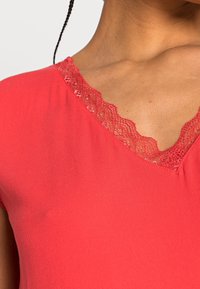 Donna che indossa un top rosso a V con rifiniture in pizzo, che mostra il collo e la parte superiore del petto.