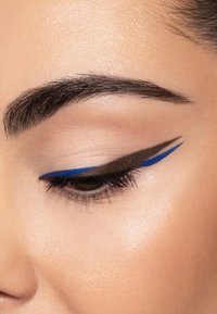 Primo piano di un occhio con eyeliner nero e blu. La pelle è liscia e leggermente tinta, con ciglia definite e una forma naturale delle sopracciglia.