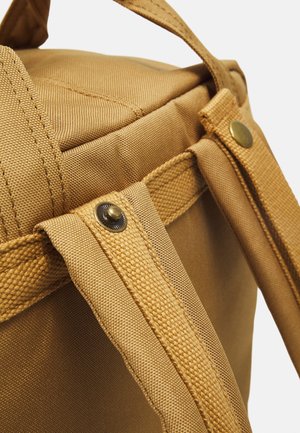 Gros plan sur des bretelles de sac à dos couleur sable avec boutons-pression en métal et détails en tissu texturé sur un fond clair.