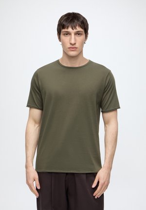 Jonge man die naar voren kijkt, draagt een olijfgroen T-shirt met korte mouwen en een donkere broek tegen een effen witte achtergrond.