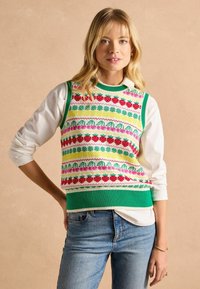 Bunt gestrickte Weste mit grünem Saum, verziert mit einem Blumenmuster in Rot-, Pink-, Gelb- und Grüntönen, darüber getragen über einem weißen Hemd.