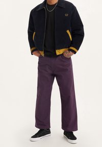 Marinblå kordjacka med gula detaljer, svart t-shirt, lila vida byxor och svarta sneakers med vita sulor. Detaljerad sömnad synlig.