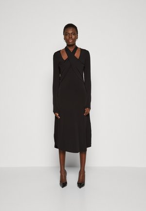 Robe noire à manches longues avec un design croisé à l'avant et une encolure dégagée, fabriquée en tissu lisse. Elle présente une longueur mi-longue et des fentes sur les côtés.