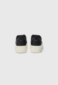 Baskets noires avec une texture lisse, une épaisse semelle blanche, et une partie arrière rembourrée. Présente un onglet de tirage à l'arrière et un design minimaliste.