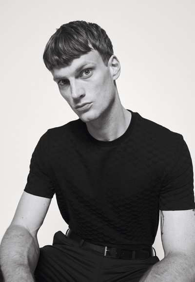 Camiseta negra texturizada con un patrón sutil, combinada con pantalones negros y un cinturón. El modelo tiene el cabello corto y está mirando directamente a la cámara.
