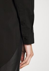 Main reposant à côté du poignet d'une chemise noire à manches longues boutonnée et d'un pantalon en cuir noir avec des coutures visibles.