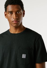 Μαύρο κοντομάνικο μπλουζάκι φτιαγμένο από μαλακό βαμβάκι με υφασμάτινη υφή, διαθέτει μπροστινή τσέπη και κωδικό της επωνυμίας προσαρτημένο.