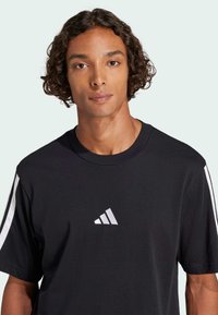 Černé bavlněné tričko s kulatým výstřihem, krátkými rukávy, bílými pruhovanými akcenty na ramenech a malým bílým logem na hrudi.