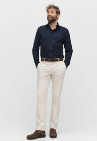 Marineblauw overhemd met knopen, lichtbeige broek, bruine riem en bruine schoenen. Model staat met handen in de zakken tegen een effen achtergrond.