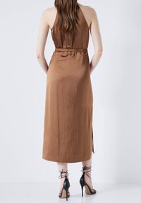 Robe sans manches marron avec une taille cintrée, tissu lisse, fentes latérales et coupe décontractée. Associée à des sandales à talons hauts noires à lanières.