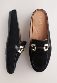 Chaussures en daim noir à enfiler avec un accent en cuir lisse, des détails en métal doré, et une semelle intérieure rembourrée beige. Design à semelle plate.