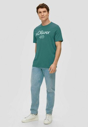 Teal cotton t-shirt with white text, paired with light blue jeans and white sneakers. Simple crew neck design, short sleeves, relaxed fit.