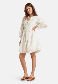 Weißes Kleid mit floralem Stickerei, V-Ausschnitt, Dreiviertelärmeln, tailliertem Bund und fließendem Rock. Getragen mit braunen Sandalen.
