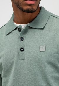 Staubgrünes Poloshirt aus weichem Stoff, mit klassischem Kragen, drei schwarzen Knöpfen und einem quadratischen Logo-Patch auf der linken Brust.
