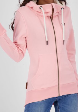 Frau, die einen hellrosa Kapuzenpullover mit Reißverschluss, weißen Kordeln und Vordertaschen trägt, kombiniert mit blauen Jeans, vor einem einfarbigen Hintergrund stehend.