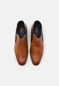 Hnědé kožené slip-on boty s hladkým povrchem a síťovanými detaily po stranách. Stélky vykazují název značky a velikost.