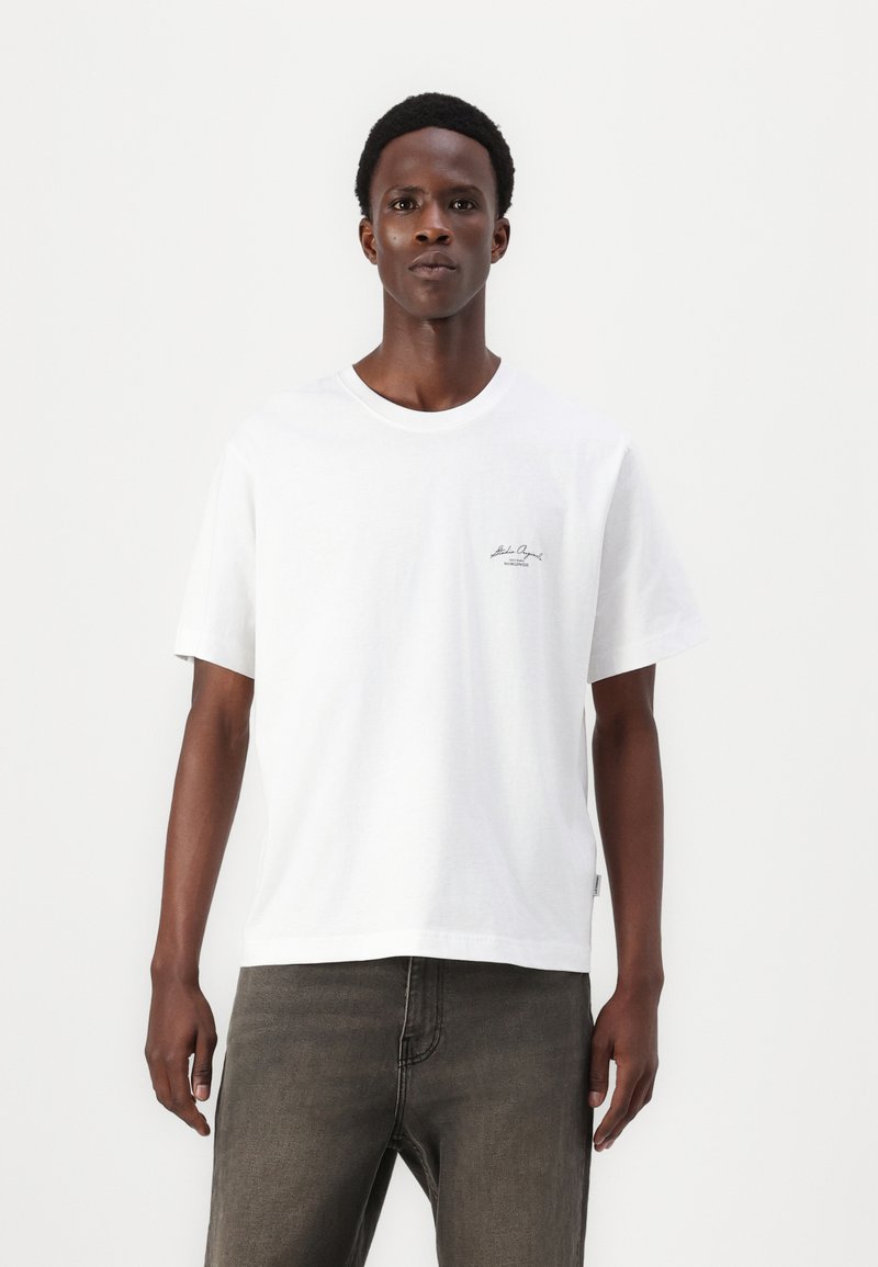 Hvit bomull t-skjorte med rund hals og korte ermer, med en liten svart logo på venstre side. Kombinert med mørkegrå jeans.