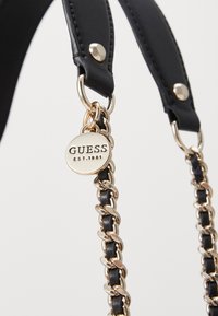 Černý popruh z umělé kůže s zlatým řetízkem a kulatou zlatou štítkem s vyrytou značkou "GUESS EST.1981." Hladký povrch s lesklým kováním.