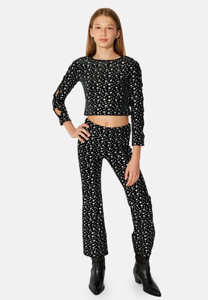 Tienermeisje staat met hand op haar heup, draagt een zwarte crop top met lange mouwen en sterpatroon, en een bijpassende broek met zwarte puntige laarzen.