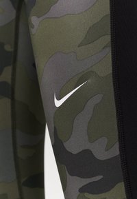 Lähikuva vihreästä ja harmaasta kamoflaažikuvioidusta kankaasta mustalla saumurilla ja pienellä valkoisella Nike-logolla.