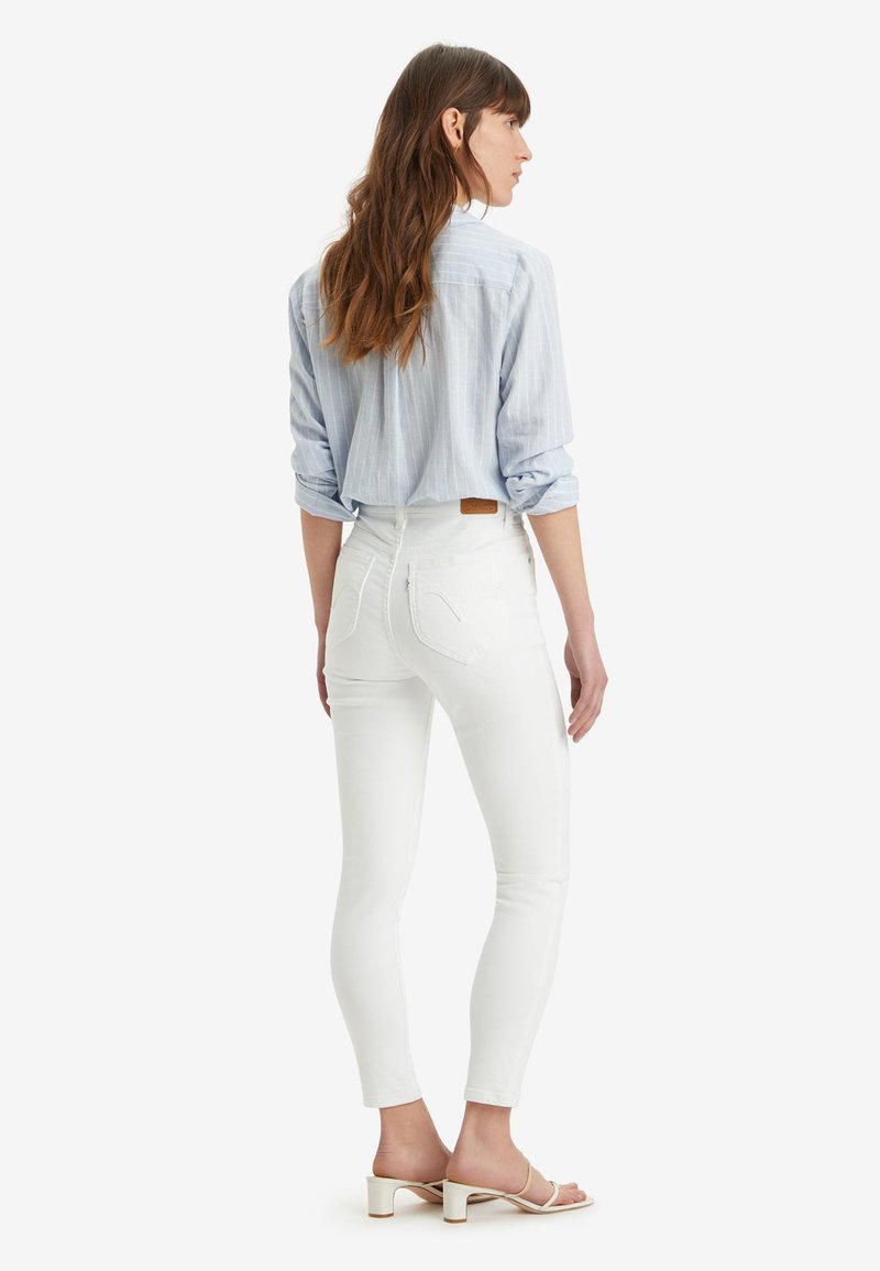 Jeans bianchi aderenti con vita alta, caratterizzati da una leggera sfumatura e dettagli di cucitura, abbinati a una camicia a righe azzurra e sandali.