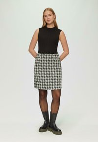Schwarzes geripptes ärmelloses Oberteil kombiniert mit einem Rock im schwarz-weißen Karomuster; das Modell trägt schwarze klobige Knöchelstiefel.