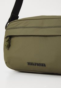 Olive green crossbody bag made of textured fabric with a zip front pocket. Features the brand logo in black and a braided zipper pull.