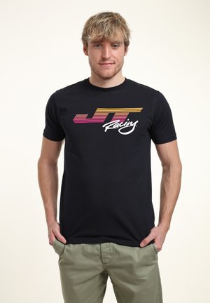 Schwarzes T-Shirt mit kurzen Ärmeln, das ein horizontales Farbverlauf-Logo in Pink, Orange und Weiß zeigt. Das Model trägt hellgrüne Hosen.