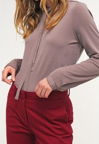 Langärmlige, mauvefarbene Bluse mit einem Vorderbinder-Detail, kombiniert mit enganliegenden, kastanienbraunen Hosen mit seitlichen Taschen und glatter Textur.