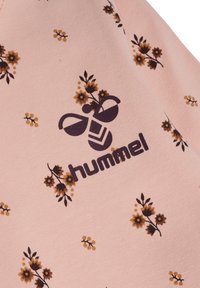 Jasnoróżowa tkanina z kwiatowym wzorem z brązowymi i żółtymi kwiatami. Logo Hummel zostało wydrukowane w ciemnym fioletowym kolorze.