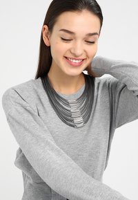 Collier en métal avec des couches de fils noirs et des accents argentés, mettant en valeur un design incurvé. Modèle portant un pull gris.