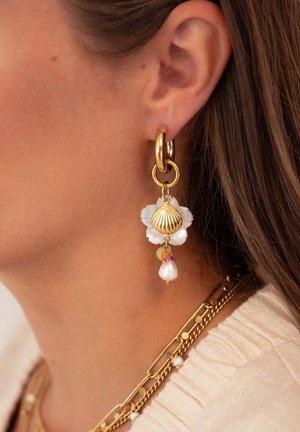 Des boucles d'oreilles en or avec un pendentif en coquillage, des accents floraux blancs et une perle pendante ornée de perles colorées, présentées sur un teint de peau.