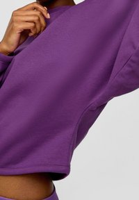 Sudadera morada con una textura suave, diseño recortado, corte relajado y cuello acanalado, mostrada parcialmente con una mano sosteniendo la tela.